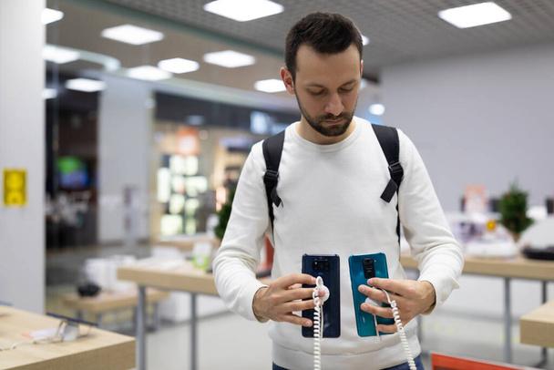 Photo of an adult handsome doubtful thinking brunette man with stubble in a white sweatshirt chooses a mobile phone in an electronic store in a shopping mall. Shop concept - Photo, Image