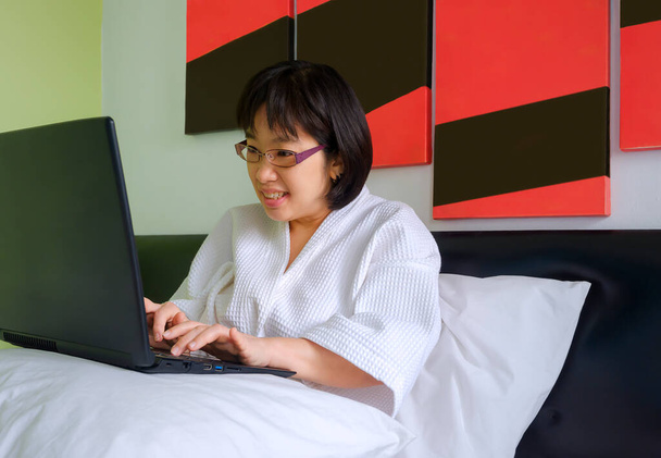 Sonriente mujer de mediana edad asiática en albornoz blanco con computadora portátil en la almohada en su cama en la mañana, acogedor concepto de relajación - Foto, Imagen