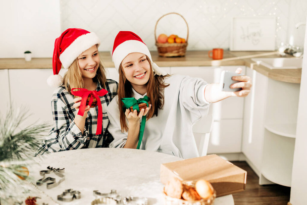 Portrait of two cute sisters in a Santa Claus Christmas hat in the kitchen, taking selfies with a smartphone, while preparing gifts on Christmas Eve and New Years Eve - Zdjęcie, obraz