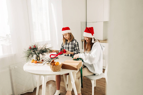 Двоє гарненьких дівчат у капелюхах Санта Клауса сидять за кухонним столом і збирають подарунки своїм друзям і родичам. Подарунок власними руками, приємне частування, святкова підготовка. - Фото, зображення