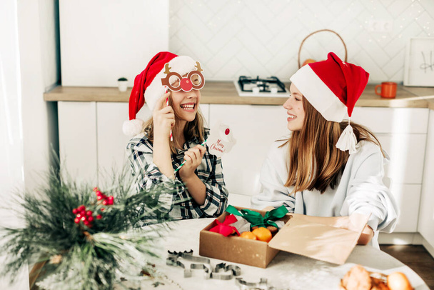 Twee meisjes in kerstmutsen pakken kerstcadeaus in, zussen hebben plezier met kerstmaskers - Foto, afbeelding