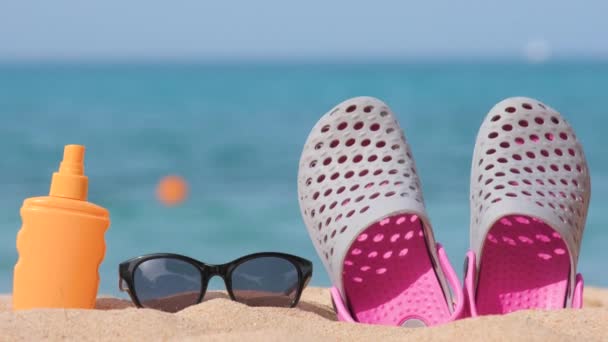 Primer plano de zapatos de zuecos, protector solar y gafas de sol protectoras negras en la playa de arena en la costa tropical en un día cálido y soleado. Concepto vacaciones de verano - Metraje, vídeo