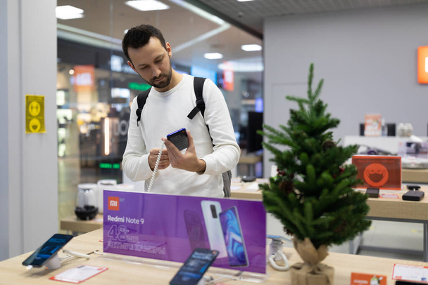 Minsk, Belarus - Nov 27, 2021: Photo of an adult handsome brunette man with stubble in a white sweatshirt chooses a mobile phone in an electronic store in a shopping mall - Valokuva, kuva