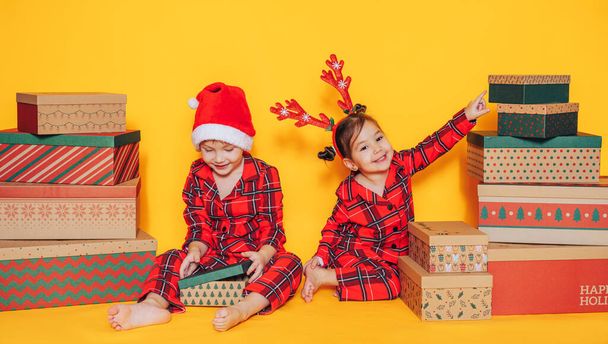 パジャマ姿の子供たちはクリスマスプレゼントと黄色い背景のオープンボックスに座っています - 写真・画像