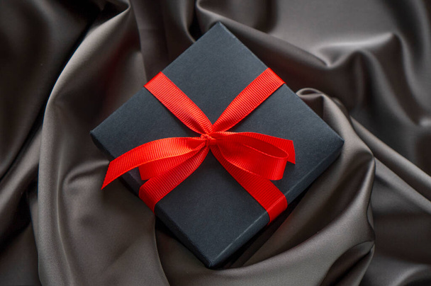 Black gift box with red bow ribbon on the satin cloth. - Photo, image
