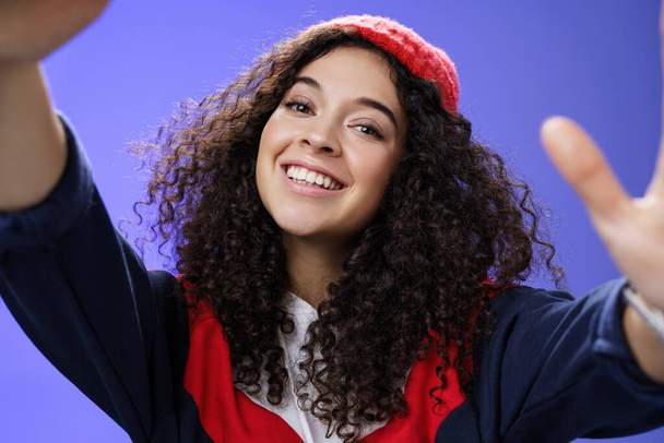 Cool and stylish friendly-looking energized happy woman in 20s with curly hair wearing warm beanie and outdoor clothes tilting head holding camera as taking selfie, smiling over blue wall - Photo, Image