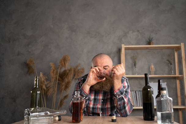adult bearded man sits at a table in casual clothes. holds a glass of whiskey in his hands and drinks. bottles of alcohol are scattered nearby. The concept of alcoholism and trying to solve problems with the help of alcohol. - Photo, Image