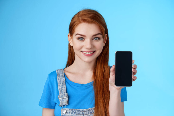 Nahaufnahme attraktive lebenslustige junge rothaarige Verkäuferin in den 20er Jahren schlägt Kunden vor, Smartphone zu kaufen, Handydisplay zu zeigen, Bewerbung zu bewerben, Internet-App, lächelnde Kamera, blauer Hintergrund - Foto, Bild