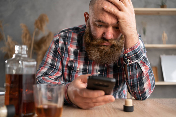 alcoholism, alcohol addiction and people concept a bearded alcoholic man with a smartphone in casual clothes sits at a table near a bottle of whiskey and a glass. Holds his head with his hand. - Photo, Image