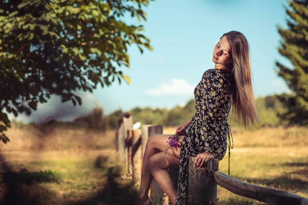 Young woman in summer dress sitting on the wooden fence - Foto, afbeelding