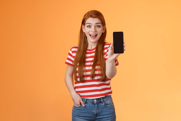 Opgewonden leuke uitgaande roodharige vrouw onder de indruk tonen app, houd smartphone introduceren gadget functie, glimlachen verrast, kijk enthousiast als toepassing, opscheppen spel score, oranje achtergrond - Foto, afbeelding