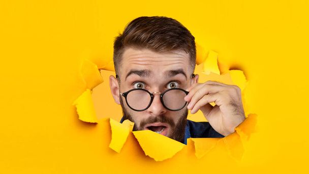 Έκπληκτος χιλιετής καυκάσιος τύπος βγάζει τα γυαλιά του και κοιτάζει στην κάμερα μέσα από την τρύπα στο κίτρινο χαρτί - Φωτογραφία, εικόνα