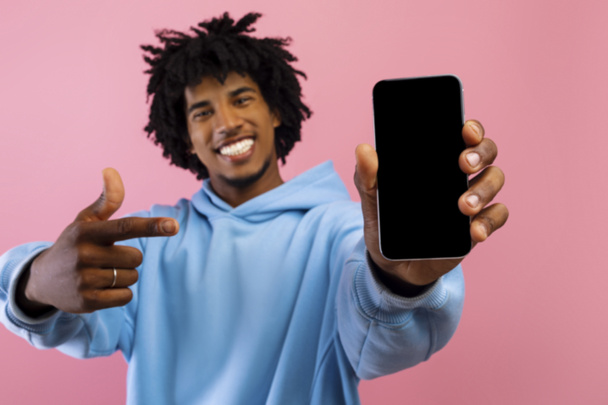 Щасливий чорний підліток вказує на смартфон з маком для веб-сайту або дизайну додатків на порожньому екрані на рожевому фоні
 - Фото, зображення