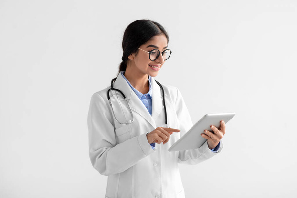 Щаслива молода індійська леді-лікар в білому пальто і окулярах, що працюють на планшеті
 - Фото, зображення