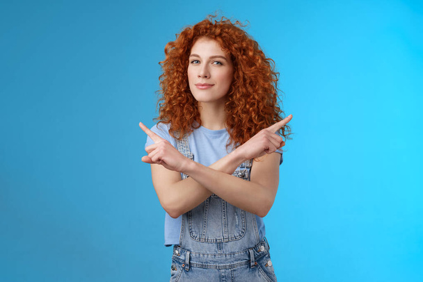 Curieux créatif rousse attrayant moderne jeune femme aux cheveux bouclés regard intrigué pointant latéralement gauche droite appareil photo qui plisse demander conseil en faisant le choix de quel produit fond bleu - Photo, image