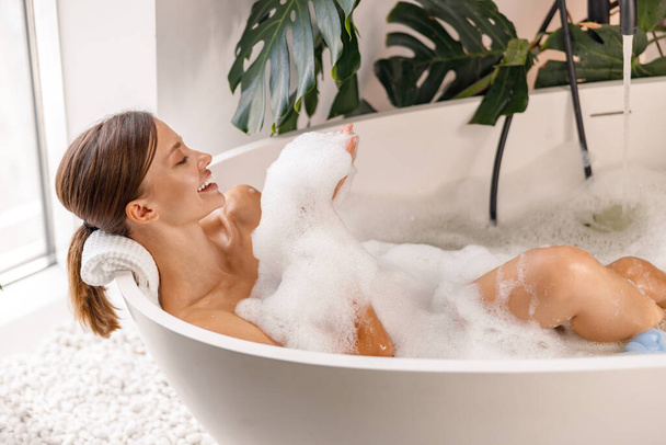 Kąpiąca się młoda kobieta relaksująca się w wannie, uśmiechnięta i bawiąca się pianką bąbelkową - Zdjęcie, obraz