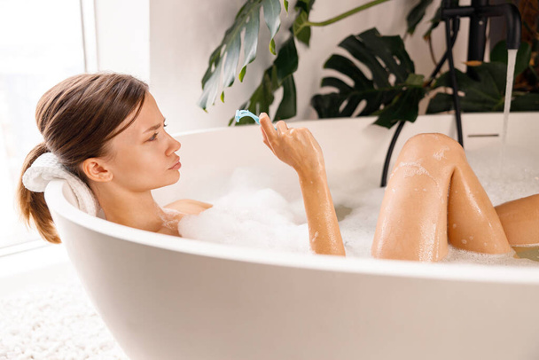 Retrato de mulher jovem fazendo caras engraçadas e segurando uma navalha para raspar o cabelo do corpo enquanto relaxa na banheira - Foto, Imagem