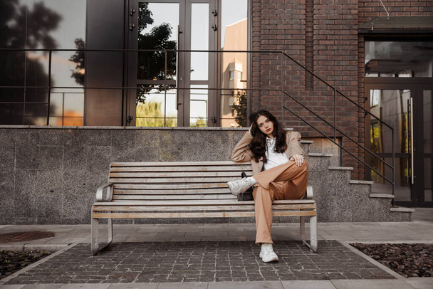  Alta chica con estilo con chaqueta de gran tamaño sentado en el banco del fondo de la ciudad - Foto, Imagen