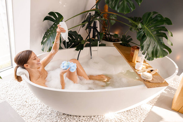 Smiling young woman pouring shower gel on loofah sponge. Wooden shelf with body care products over bubble filled bathtub - Φωτογραφία, εικόνα