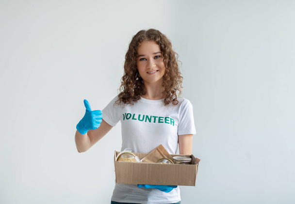 Excited activist volunteer holding box with donations and showing thumb up, smiling at camera over light background - Photo, Image