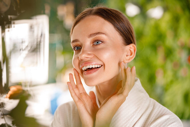 Крупный план портрета великолепной молодой женщины с гладкой кожей, улыбающейся во время процедуры красоты в ванной комнате - Фото, изображение