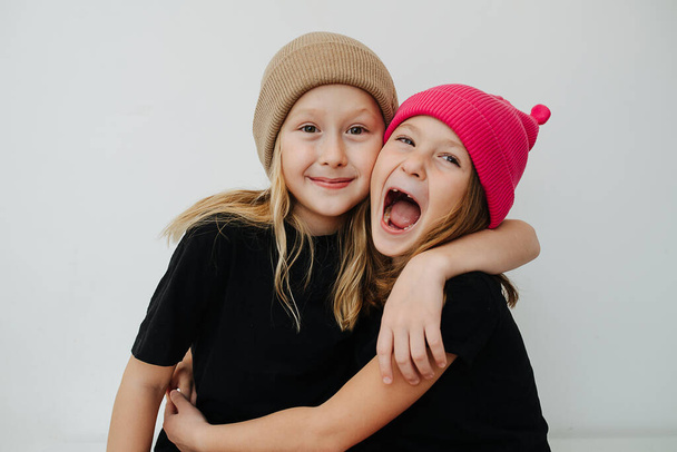 Friendly siblings in colorful knitted hats hugging, touching their cheeks. Over white wall background. - Foto, imagen