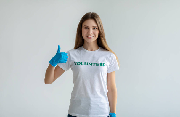 I like volunteering. Positive lady in white volunteer t-shirt showing thumb up, wearing blue gloves, light background - Photo, Image