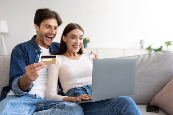 Smějící se mladý evropský pár na pohovce s kreditní kartou, notebookem a nakupováním online v obývacím pokoji - Fotografie, Obrázek