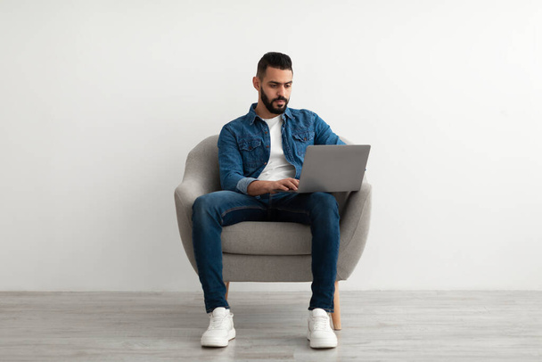 Longitud completa del hombre árabe usando el ordenador portátil para el trabajo en línea, los estudios o la comunicación, sentado en sillón, pared blanca del estudio - Foto, imagen