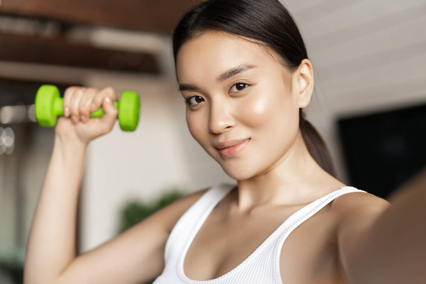 Retrato de sonriente asiática instructor de fitness tomando selfie con mancuerna. Mujer atlética haciendo foto en el teléfono móvil con equipo deportivo, entrenamiento en casa - Foto, imagen