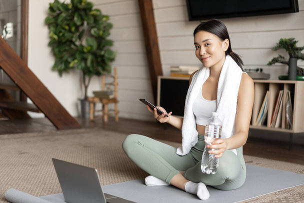 Asian girl in fitness clothing sitting at home on floor mat, resting after workout, checking mobile phone sport application - Foto, Imagem