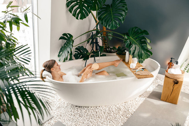 Uvolněná mladá žena pomocí štětce při koupeli v bublině v moderní koupelně zdobené tropickými rostlinami - Fotografie, Obrázek