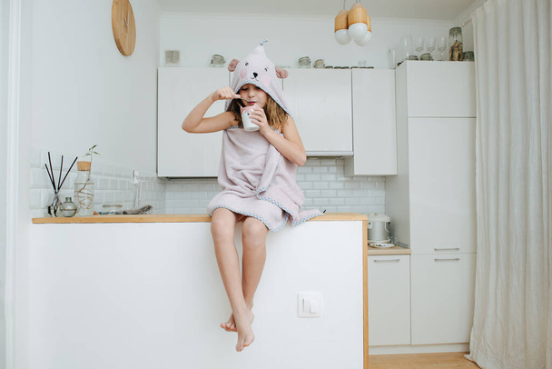 Focused on eating little girl wrapped in a bear hooded towel. She is sitting on a high table, having yogurt for a supper in a kitchen. - Foto, Bild