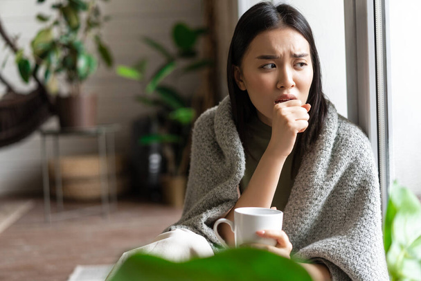 Üzgün Asyalı kız kendini karantinaya almış, grip olmuş ve elinde bir bardak çayla evde oturmuş öksürüyor. - Fotoğraf, Görsel