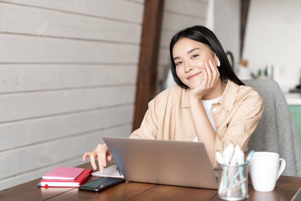 Uśmiechnięta Azjatka pracująca zdalnie z domu, siedząca z laptopem i telefonem komórkowym. Koreańska dziewczyna odrabia lekcje, e-learning z komputerem z kuchni - Zdjęcie, obraz