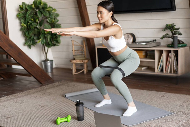 Schattig aziatisch fitness meisje thuis doen workout, hurken met stretching elastische touw op benen, staan op vloermat, luisteren naar online sport instructeur op laptop video - Foto, afbeelding