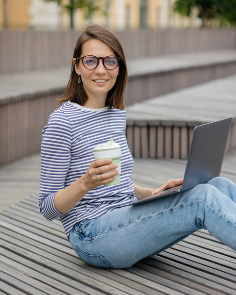 young and confident woman blogger or marketer drinking coffee and working on her laptop. work in a park or public place - Foto, Imagen