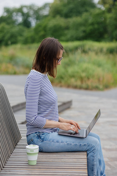 young and confident woman blogger or marketer drinking coffee and working on her laptop. work in a park or public place. social media or work as a top manager. - 写真・画像