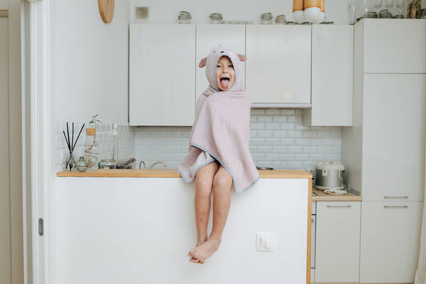 Funny little girl wrapped in a bear hooded towel sitting on a high kitchen table. She's sticking her tongue out, long shot. - 写真・画像