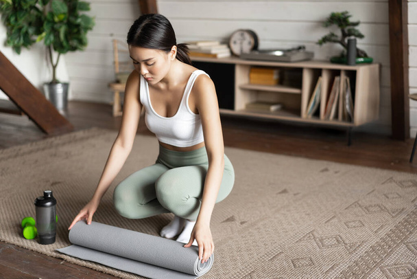 Asiático fitness girl terminar o treinamento, treino em casa, tapete de rolamento após o exercício na sala de estar, limpeza - Foto, Imagem