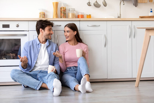 Wesoły uśmiech młodej żony i męża z filiżanek gorącego napoju siedzi na podłodze i rozmawia w kuchni - Zdjęcie, obraz