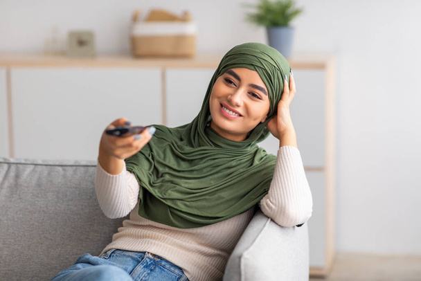 Fiatal arab nő muszlim hidzsábban távirányítóval a kezében, otthon tévét néz a kanapén - Fotó, kép
