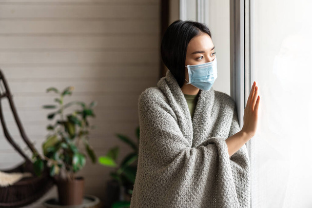 Beteg ázsiai lány orvosi maszkban áll az ablaknál, és arra vágyik, hogy kimehessen, karanténban van, beteg, vagy influenzás. - Fotó, kép