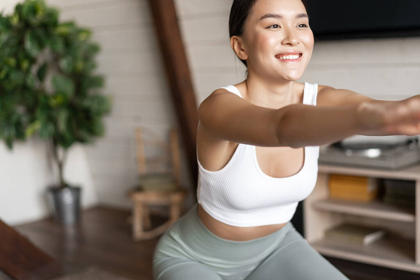 Sağlıklı vücut yapısı olan genç Asyalı fitness kadını, çömelme hareketleri, sabah antrenmanı, aktif giyimi, oturma odasında ayakta durmak ve antrenman yapmak. - Fotoğraf, Görsel
