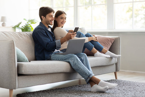 Frohe fröhliche Millennial-Europäer sitzen mit Laptop auf dem Sofa und schauen aufs Smartphone - Foto, Bild