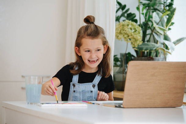 Lively little girl sitting behing a big table next to a window, painting in front of a laptop, using it as a template. She is showing her missing teeth. - Photo, image