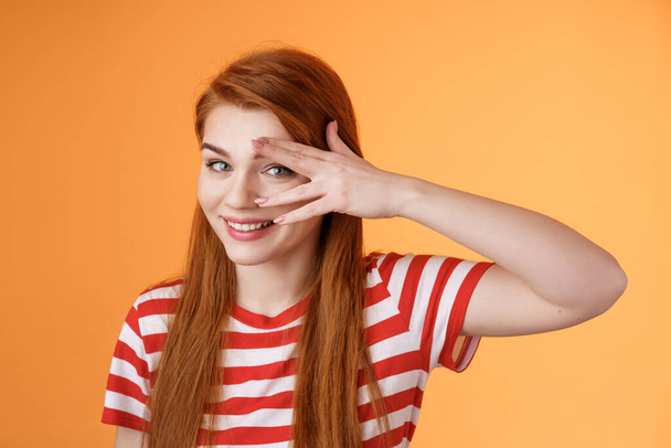 Charismatic lovely redhead lucky female, smiling cute, peek camera tender coquettish gaze, hold palm face, look through fingers, stand orange background optimistic, cheerful mood - Photo, Image
