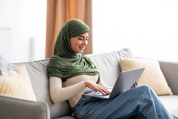 若いアラブ人女性の肖像画ヒジャーブのソファに座って、ラップトップを使用して、ウェブサーフィン、自宅でリモートで仕事や勉強 - 写真・画像