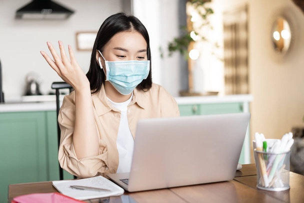 Mujer asiática alegre trabajando desde casa, comunicándose en el ordenador portátil, usando máscara médica. Chica en cuarentena estudiando a través de clases en línea - Foto, Imagen
