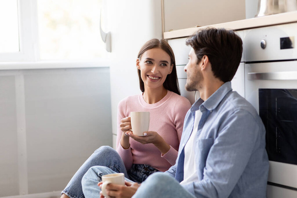 Satisfied smiling young lady and man with cups of hot drink sits on floor, flirting and talking at kitchen interior - Фото, изображение